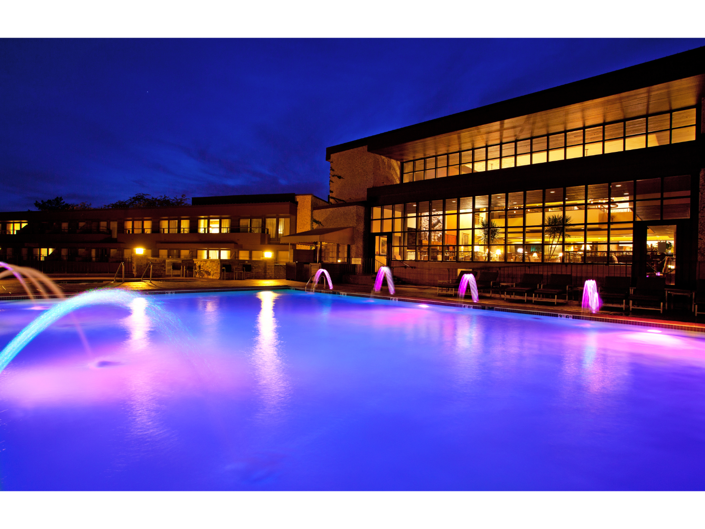 Evening view of a resort's illuminated pool area, featuring colorful lights reflecting off the water.