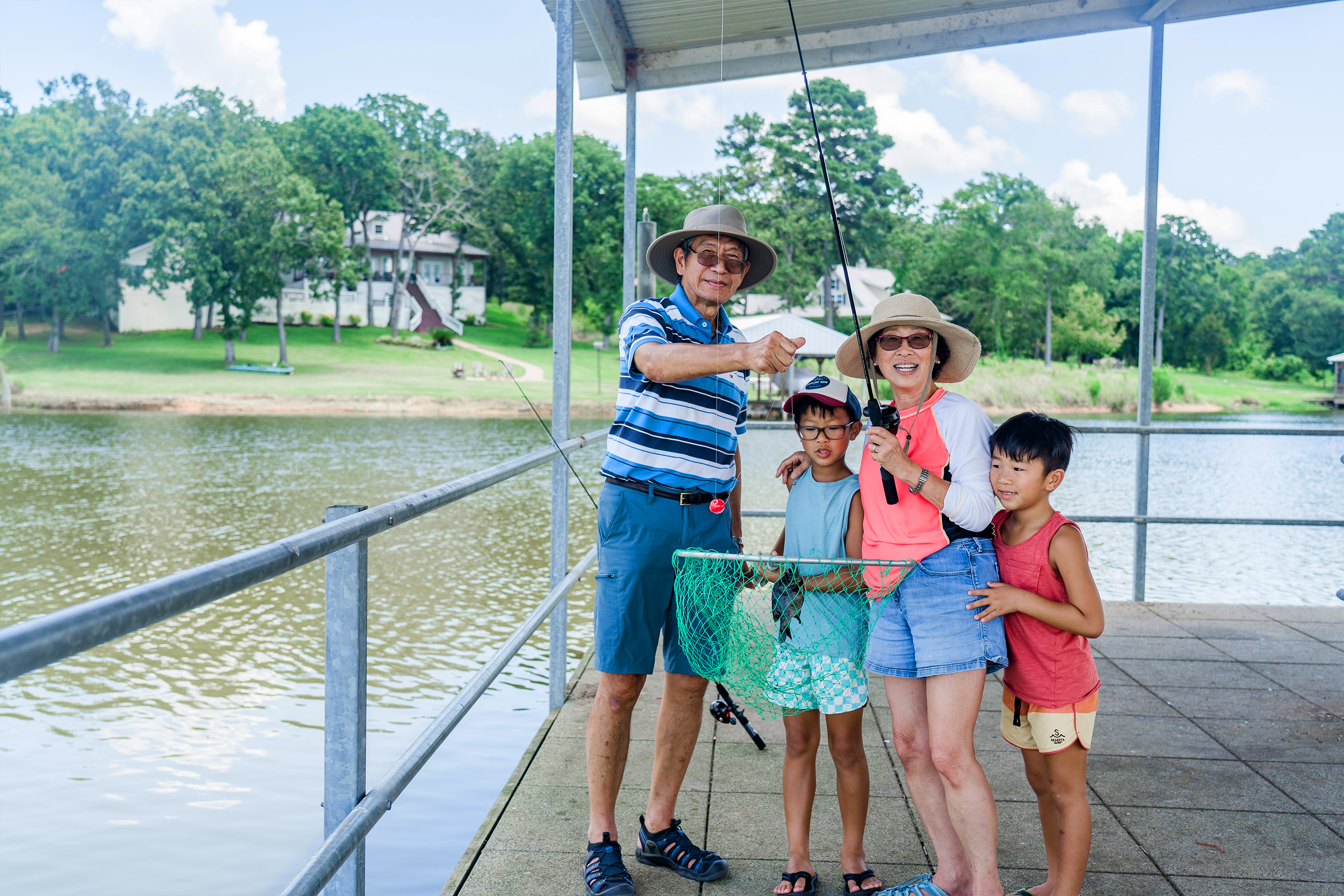 An older Asian male (left) and female (middle-right) surrounded by two young male boys wear  sunglasses and various hats while holding up fishing gear and their catch.