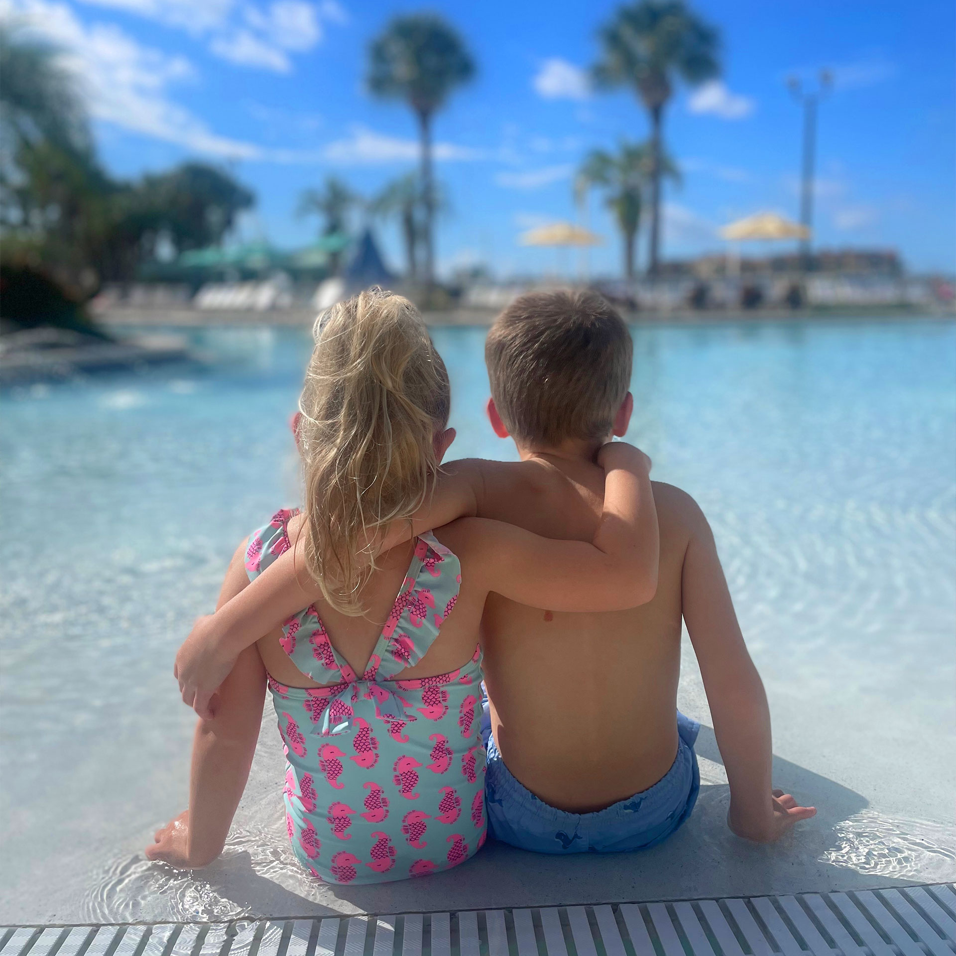 A boy and girl wits holding arms around each other in swimsuits near the edge of an outdoor, zero-entry pool.