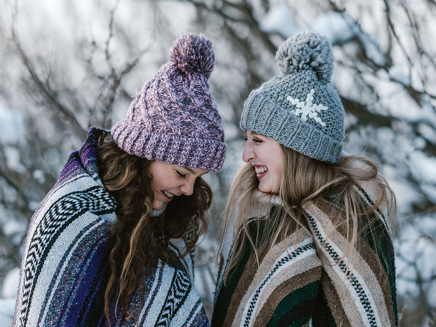 Two girls outside in the snow wearing blankets and beanies while laughing.