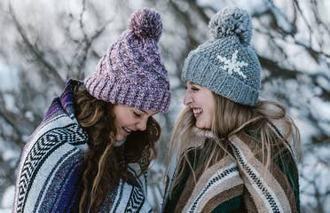 Two girls outside in the snow wearing blankets and beanies while laughing.