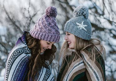 Two girls outside in the snow wearing blankets and beanies while laughing.
