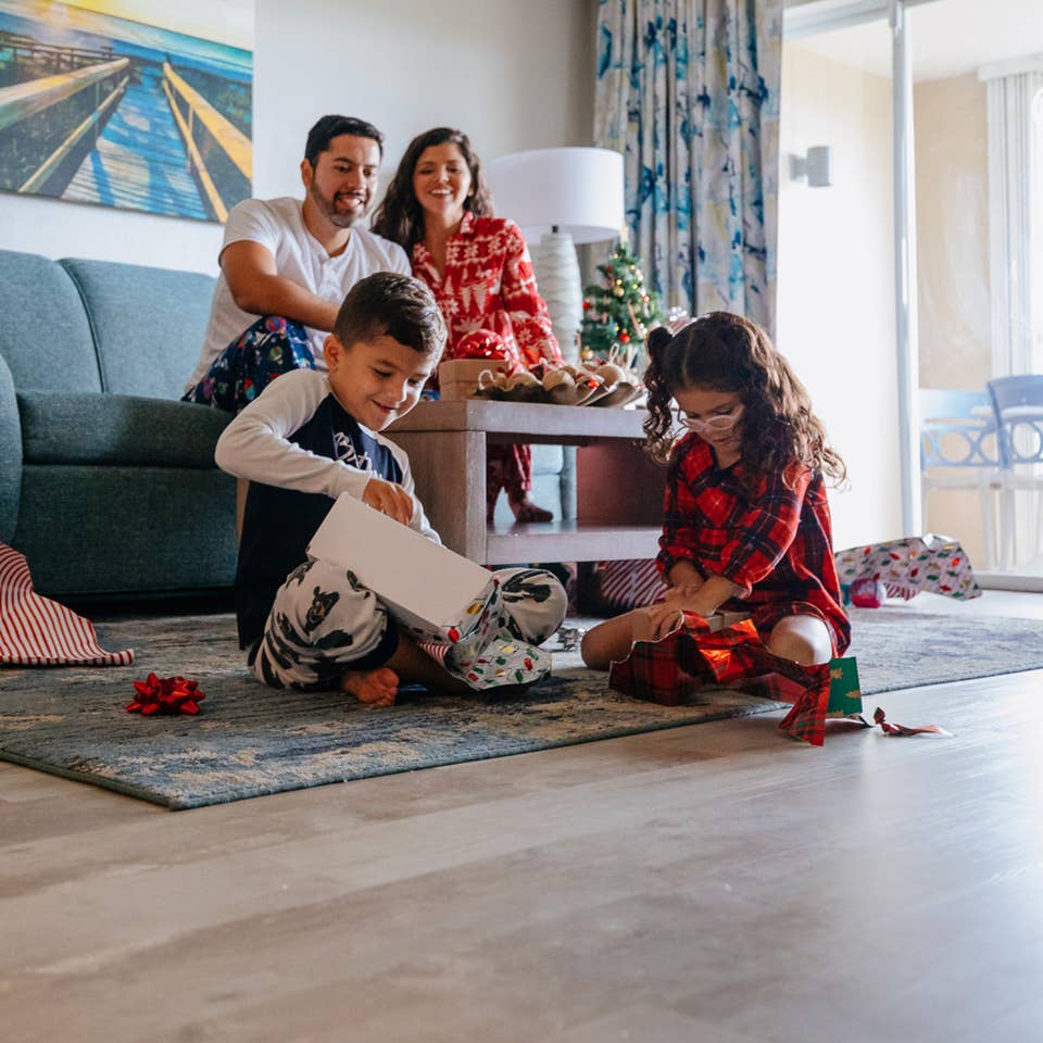 A man, young boy and woman watch a young girl unwrap a present in a living room while all wearing pajamas.