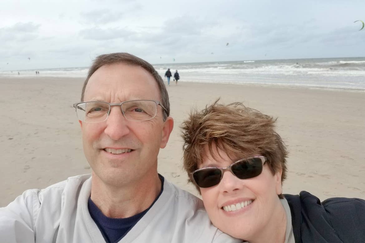 A caucasian man (left) wears a light grey crewneck sweater next to a caucasian woman (right) on a beach in Zandvoort, Netherlands.