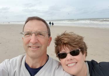 A man (left) wears a light grey crewneck sweater next to a woman (right) on a beach in Zandvoort, Netherlands.