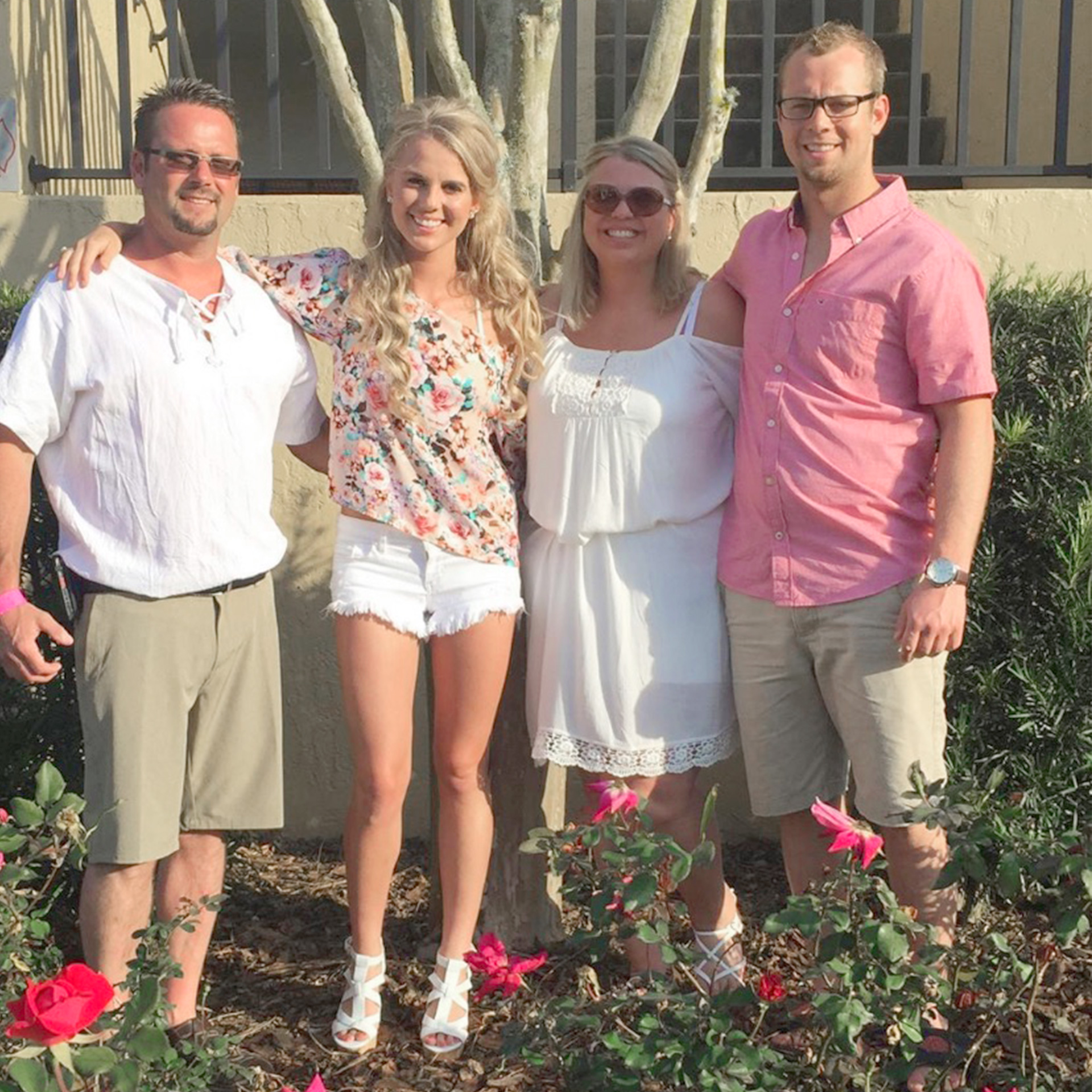 A caucasian male (left), two Caucasian females (middle) and another Caucasian male (right) stand in front of a stucco staircase behind some red rose bushes.