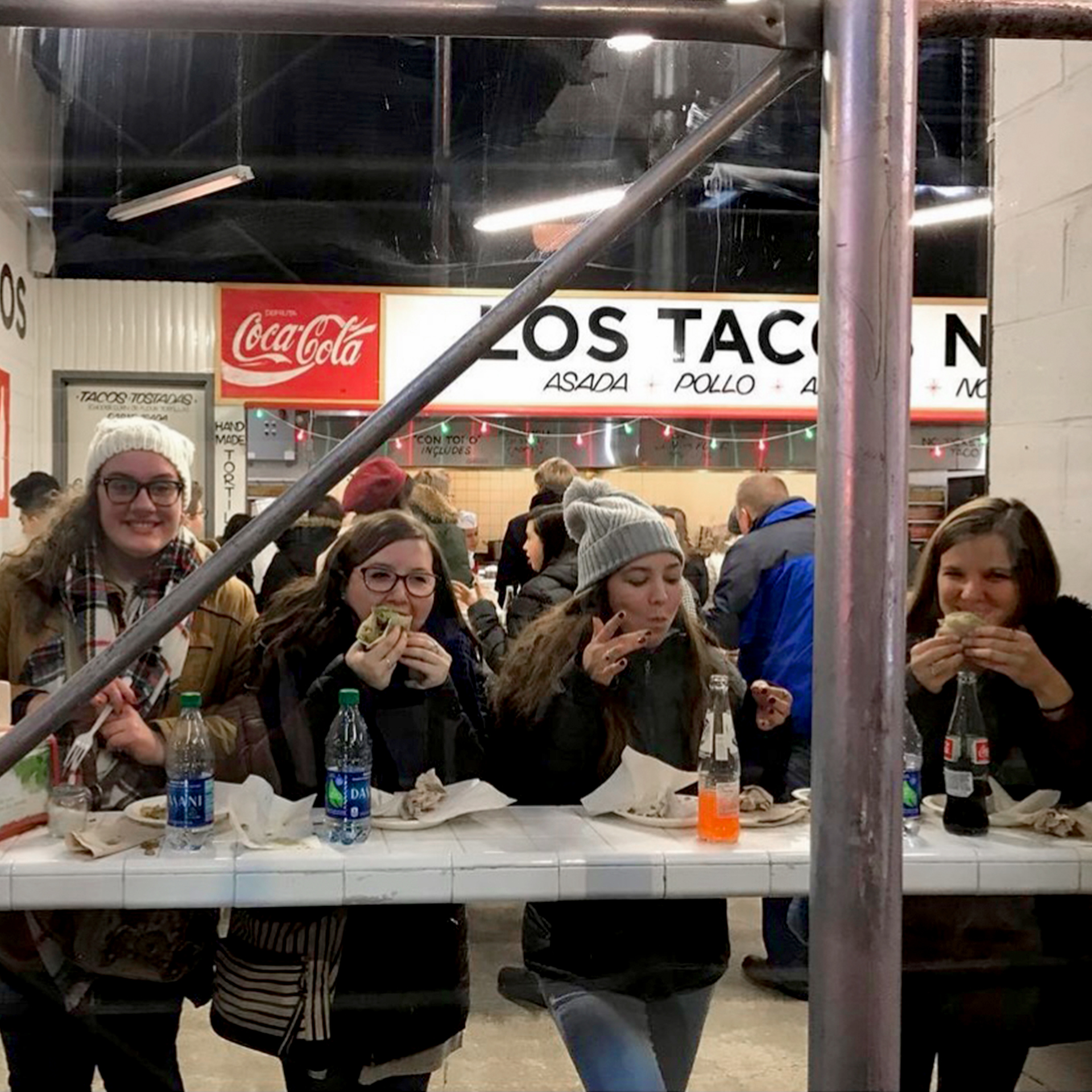 Four caucasian women wearing winter apparel are seated at a countertop at 'Los Tacos' in New York City.