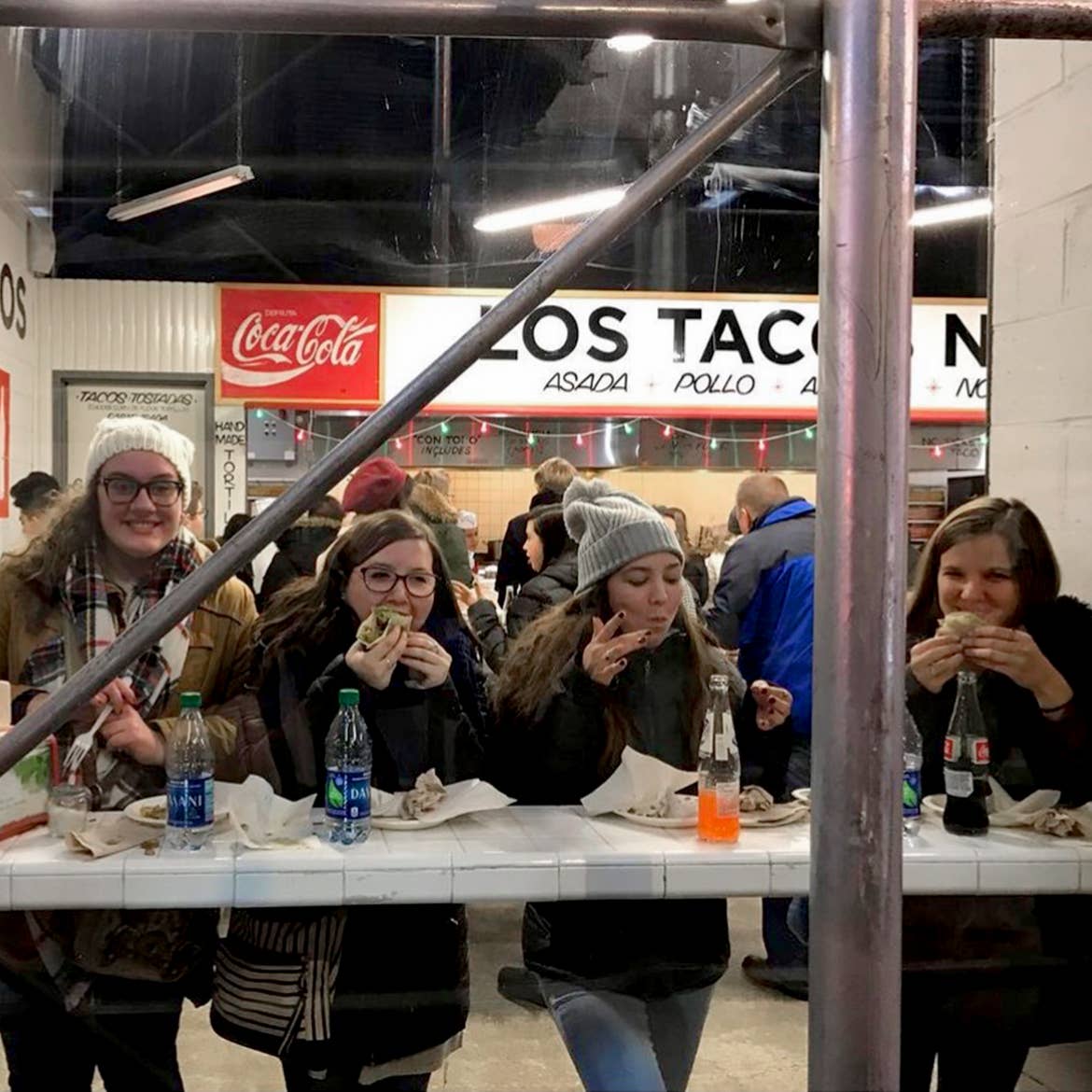Four caucasian women wearing winter apparel are seated at a countertop at 'Los Tacos' in New York City.