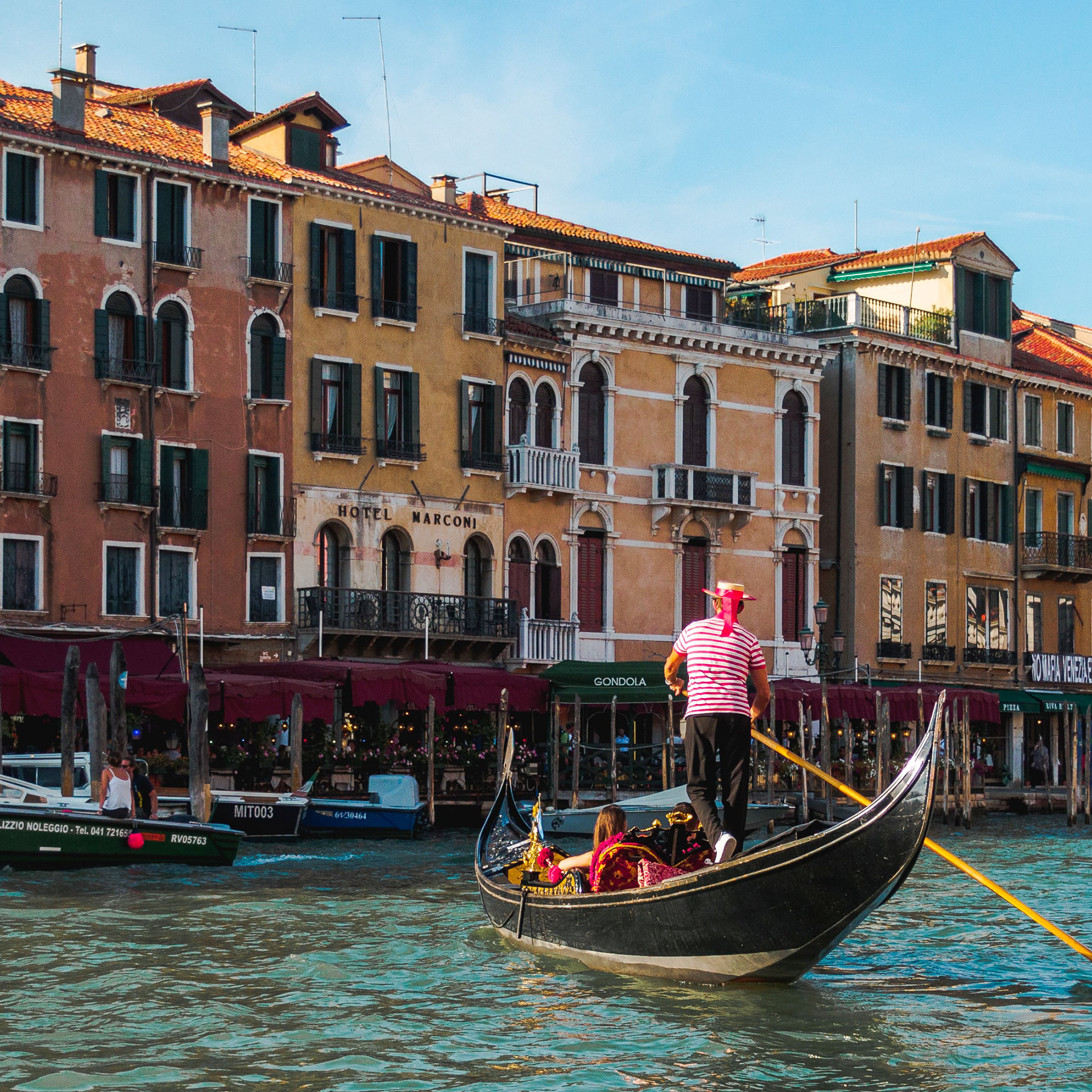 A Gondolier wearing a straw hat and striped red and white shirt paddles guests along the river in Venice, Italy.