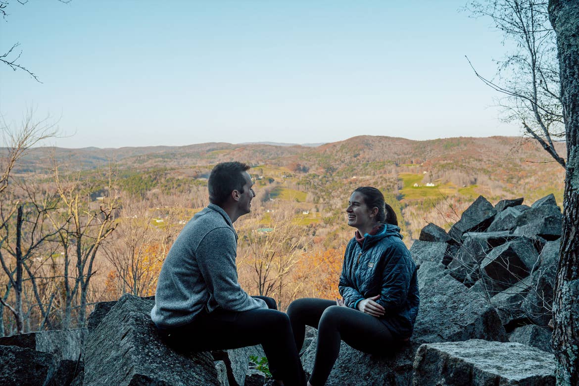 A man (left) and woman (right) wearing sweaters and pullovers with black pants sit on rock formations near the edge of a hill overlooking trees with fall foliage.