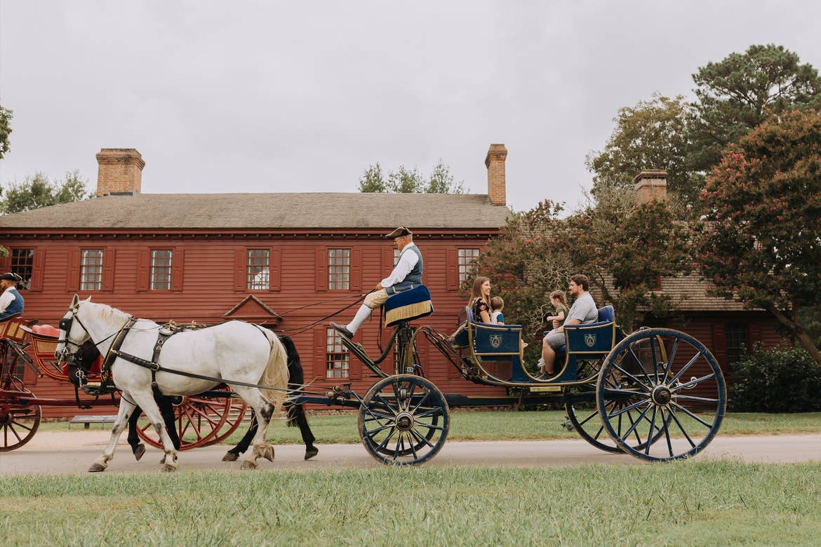 A family sits on a horse-drawn carriage driven by an actor wearing period-appropriate attire at Colonial Williamsburg in Williamsburg, Virginia.