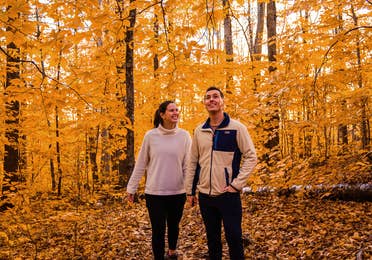 A woman (left) and man (right) wearing white sweaters and pullovers with black pants stand in the middle of trees with yellow fall foliage.