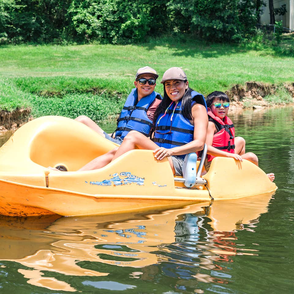 An Asian Male (left), female (middle) and young child (right) wear blue life vests, sunglasses and various hats while riding on a yellow paddleboat in a lake.