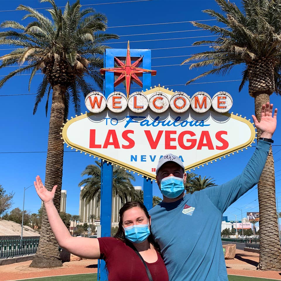 Featured Contributor, Ashley Fraboni (left) and her fiancé, Nicholas (right), pose in front of the 'Welcome to Las Vegas' marquee while wearing face masks.