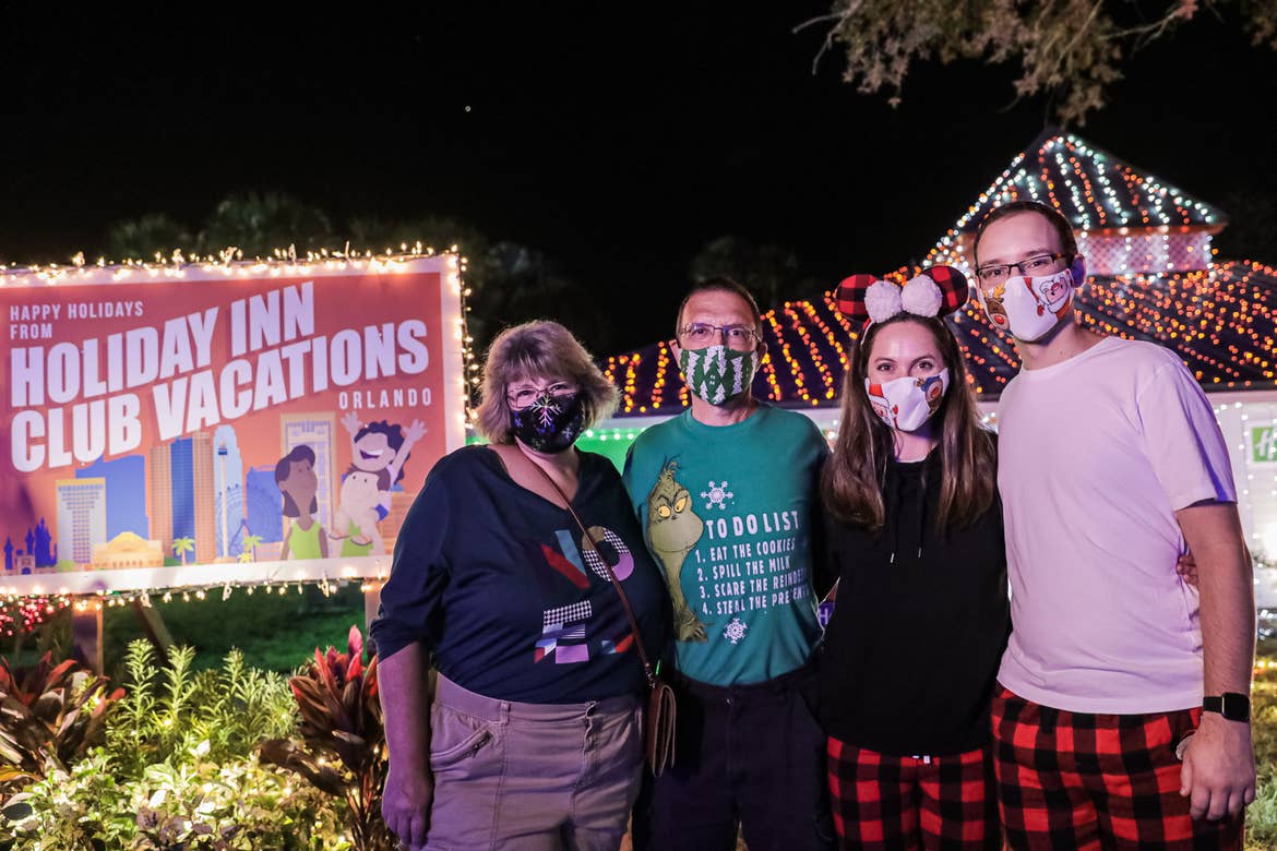 Two women and men stand in front of a building exterior decorated with multi-colored string lights while wearing safety masks.