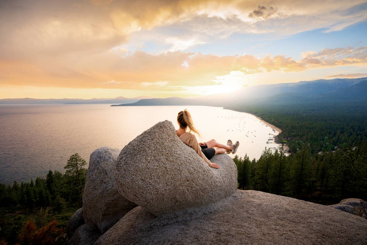 A caucasian woman wearing a beige sweater, black biker shorts and boots sits on a rock formation at a high elevation overlooking a lake surrounded by pine trees on a rocky shoreline.