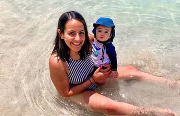 A woman in a striped swimsuit holds a baby wearing swimsuit and a sunhat in shallow water.