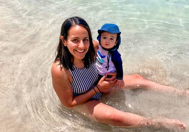 A woman in a striped swimsuit holds a baby wearing swimsuit and a sunhat in shallow water.
