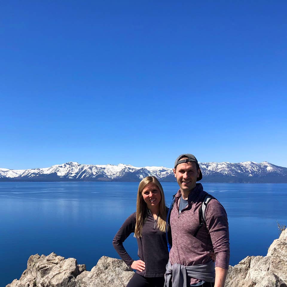 A woman (left) black pullover while hugging a man (right) wearing a red pullover and grey hat in front of a mountain range at Lake Tahoe, Nevada.