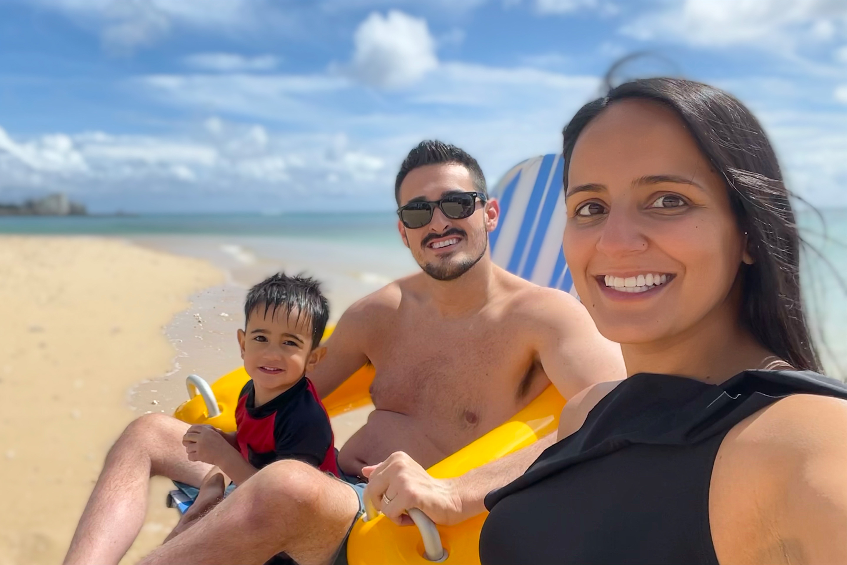 Featured Contributor, Danny Pitaluga (middle), sits in a yellow beach wheelchair on the sand as waves roll in while holding Joey (left) while his wife, Val (right) sits offside.