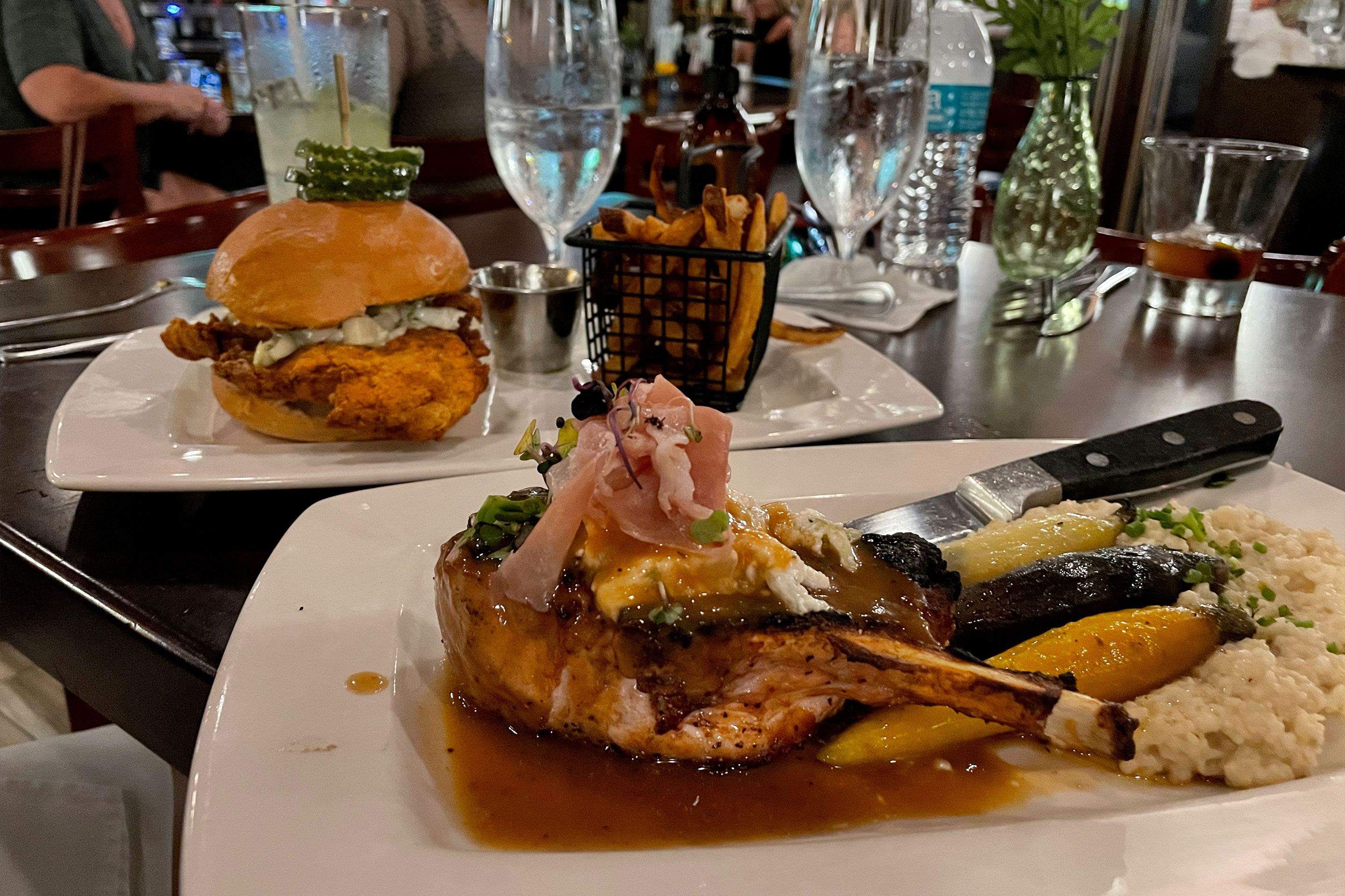 Two white, plated dishes containing a sandwich (back) and pork chop (front) placed on a dark tabletop.