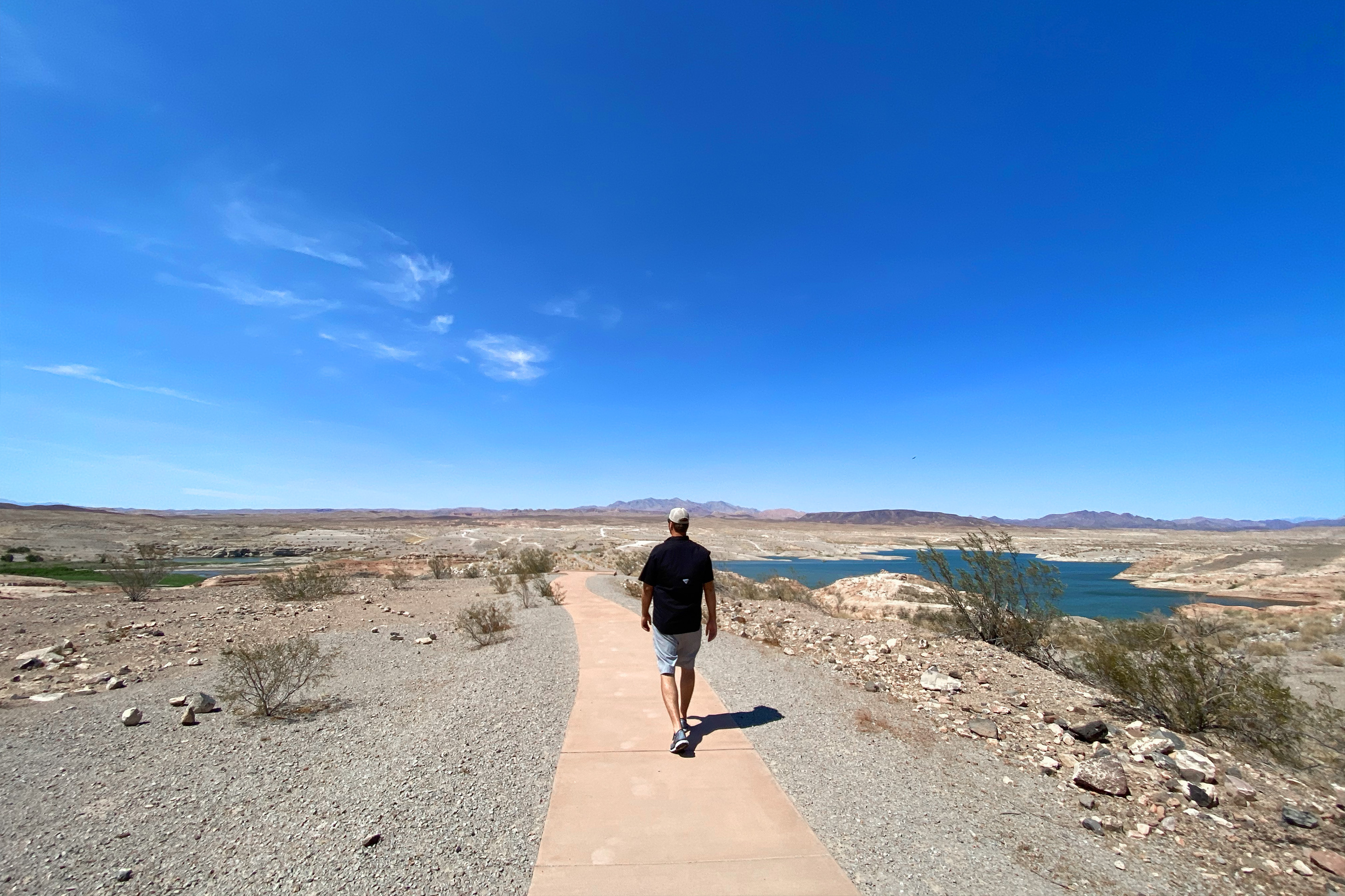 A man wearing a white hat, black t-shirt, and denim shorts walks on a path at the Lake Mead National Recreation Area under a blue, lightly-clouded sky.