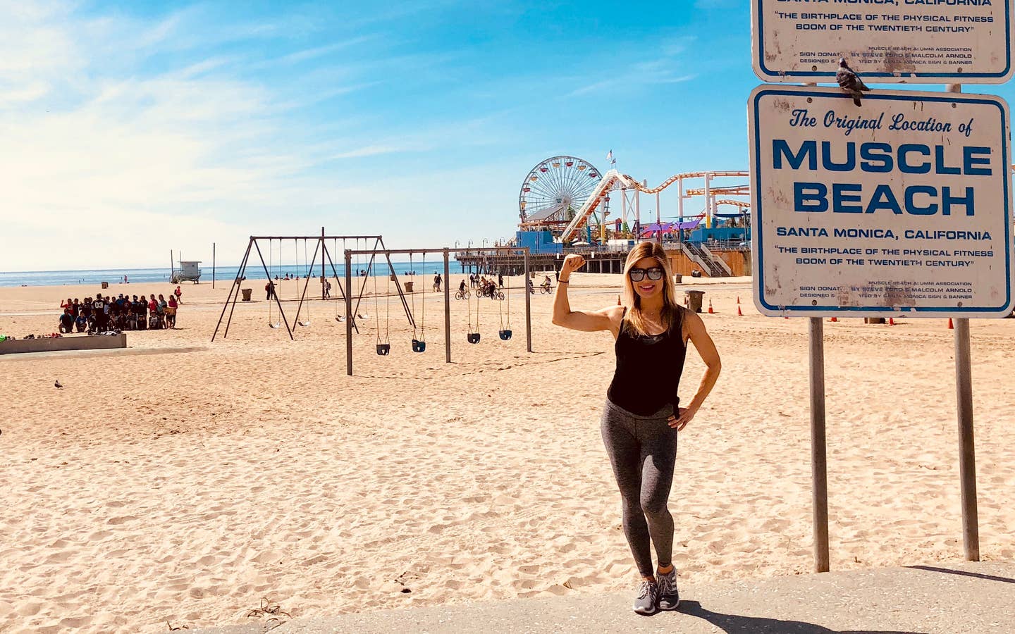 Featured Co-author, Christine, stands on Venice Beach wearing black tanks and leggings in front of the boardwalk and various exercise areas.