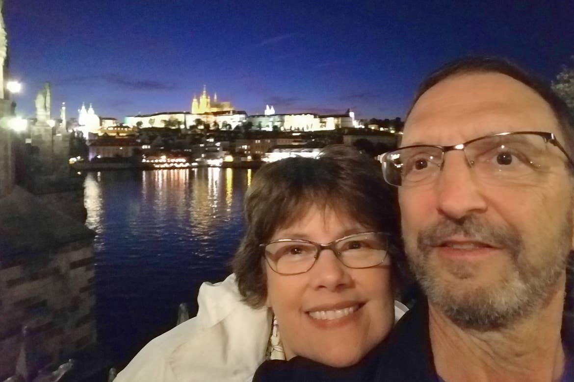 A caucasian male (right) wearing a navy shirt and a caucasian woman (left) wearing a white windbreaker jacket stand at a distance from the city of Prague illuminated under the night sky.