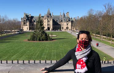 Author, Jenn C. Harmon, stands in front of the Biltmore Estate wearing a buffalo plaid red and black face mask and festive scarf over a black jacket.