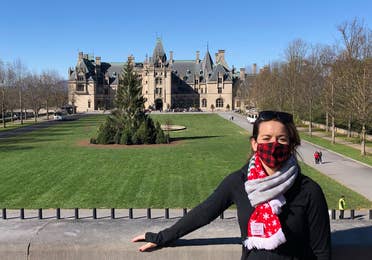 Author, Jenn C. Harmon, stands in front of the Biltmore Estate wearing a buffalo plaid red and black face mask and festive scarf over a black jacket.