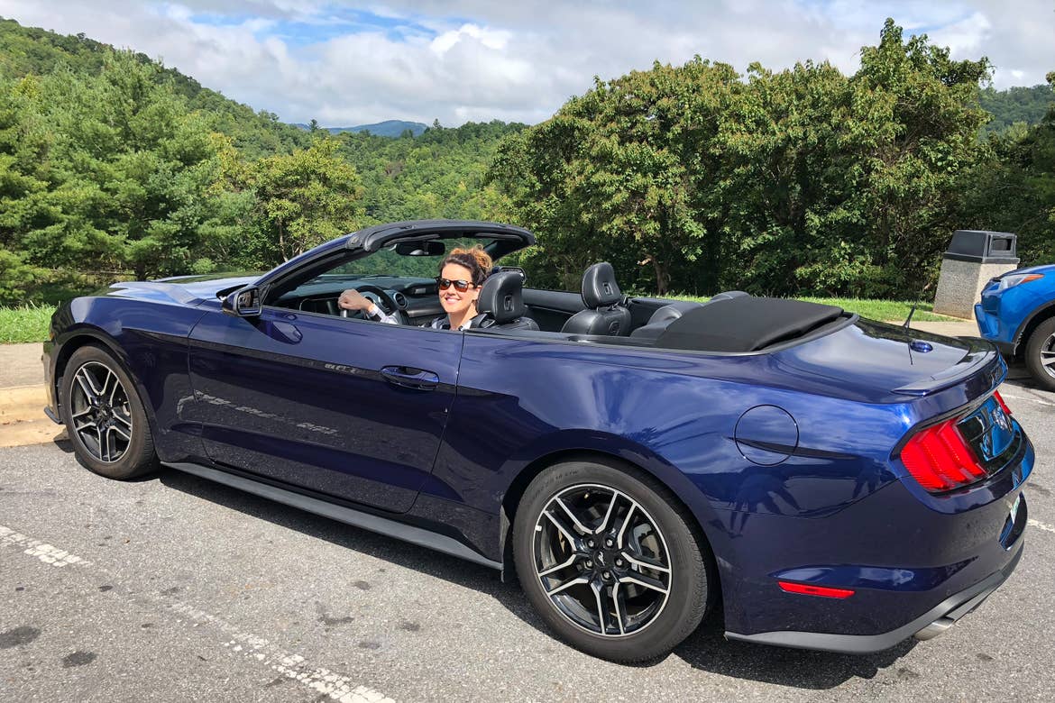 Jenn C. Harmon sits in the driver's seat of a dark blue Mustang convertible surrounded by lush foliage near the Great Smoky Mountains.