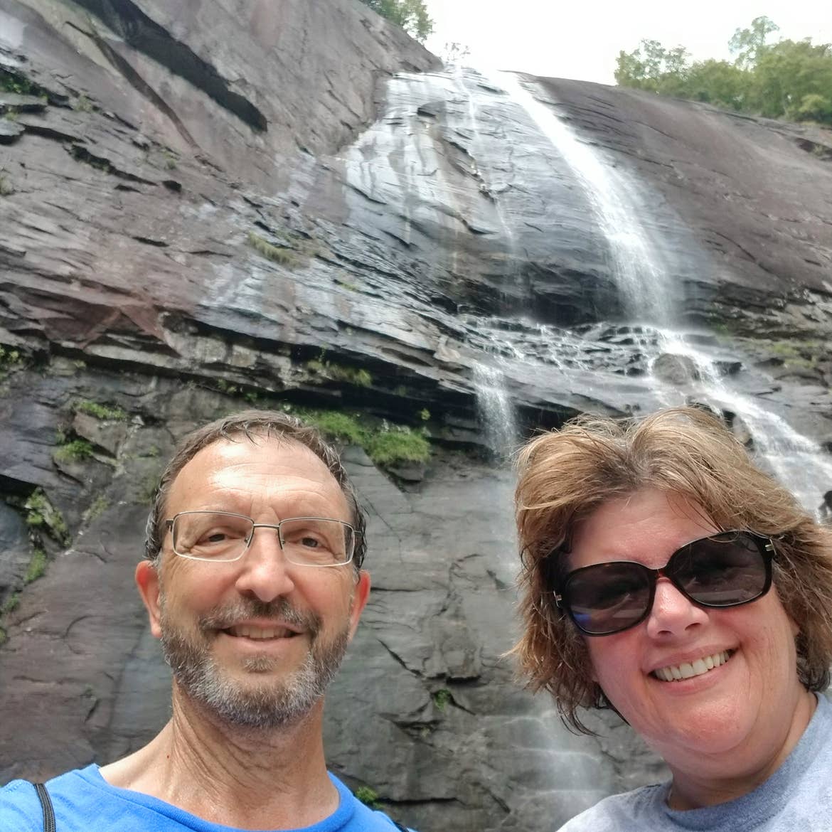 A caucasian male wearing a blue t-shirt (left) and a caucasian female wearing a light colored t-shirt (right) stand under the Hickory Nut Falls in Chimney Rock State Park, North Carolina.