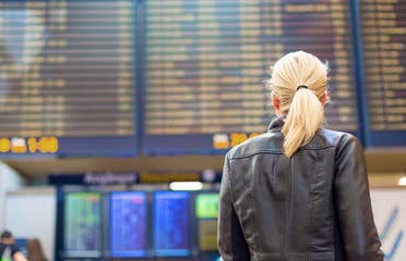 A blonde woman faces an airport Departure board in the terminal while wearing a black leather jacket.