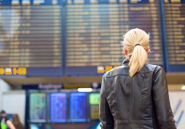 A blonde woman faces an airport Departure board in the terminal while wearing a black leather jacket.