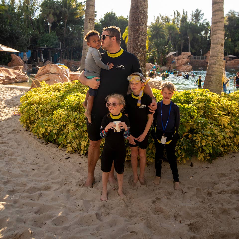 A man and four children wear black and yellow wetsuits on a sandy beach under the sun and pal trees.