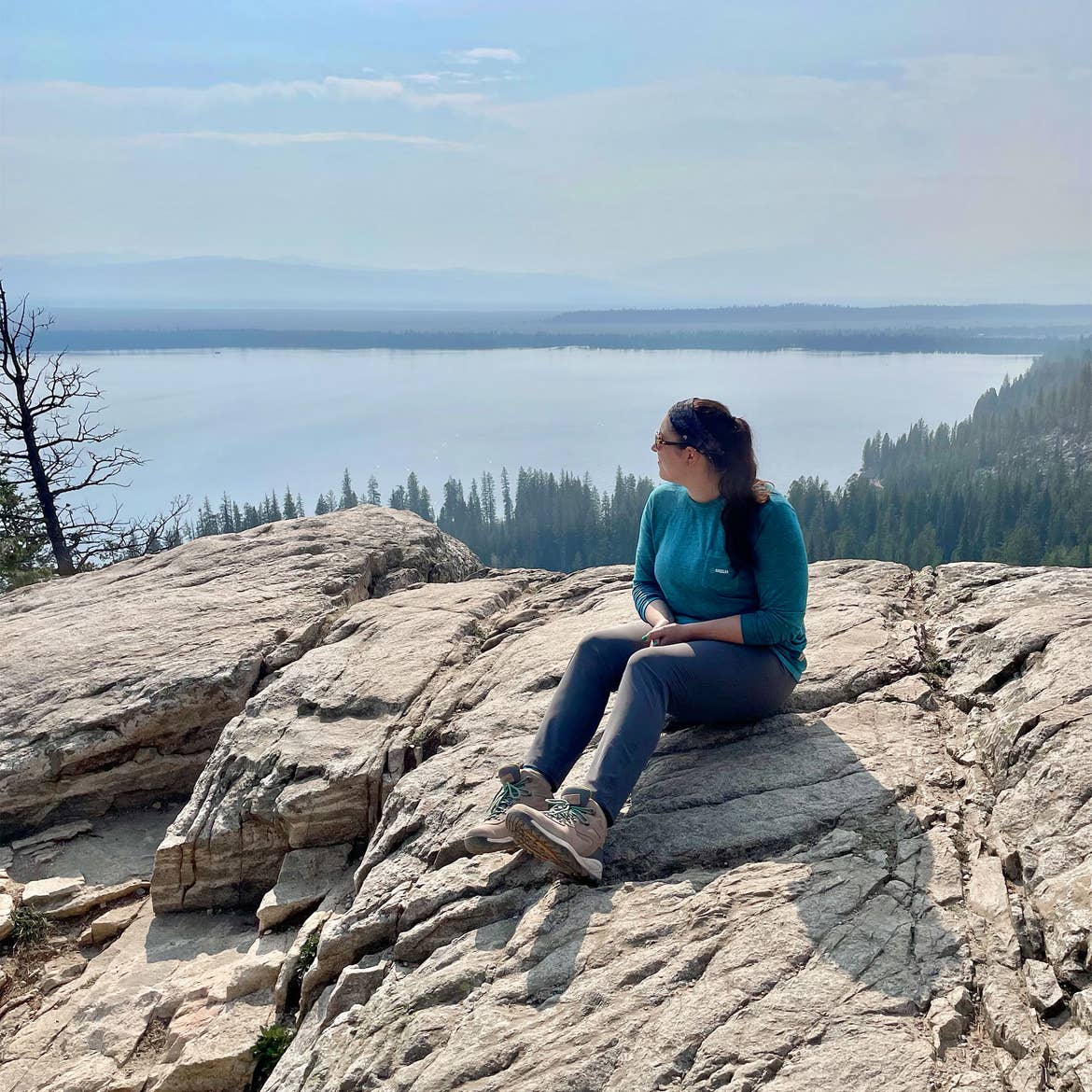 A woman in a blue, long sleeve shirt, grey pants, and brown hiking boots sits on a rock formation on a mountain overlooking a lake.