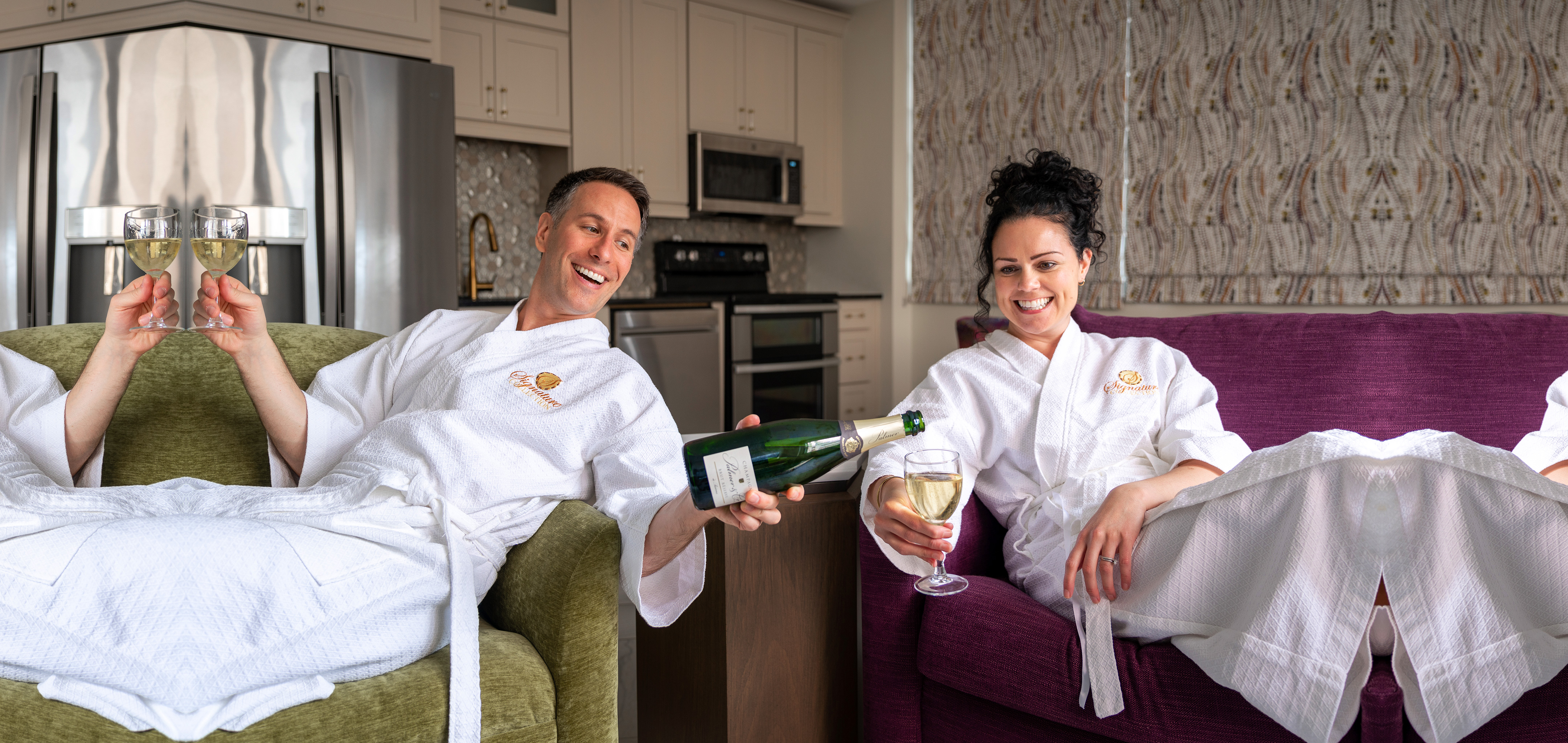 A man wearing a white robe on a green couch (left) holds a glass of champagne in his right hand while pouring the green bottle in this left hand for a woman (right) also wearing a white robe on a purple couch.