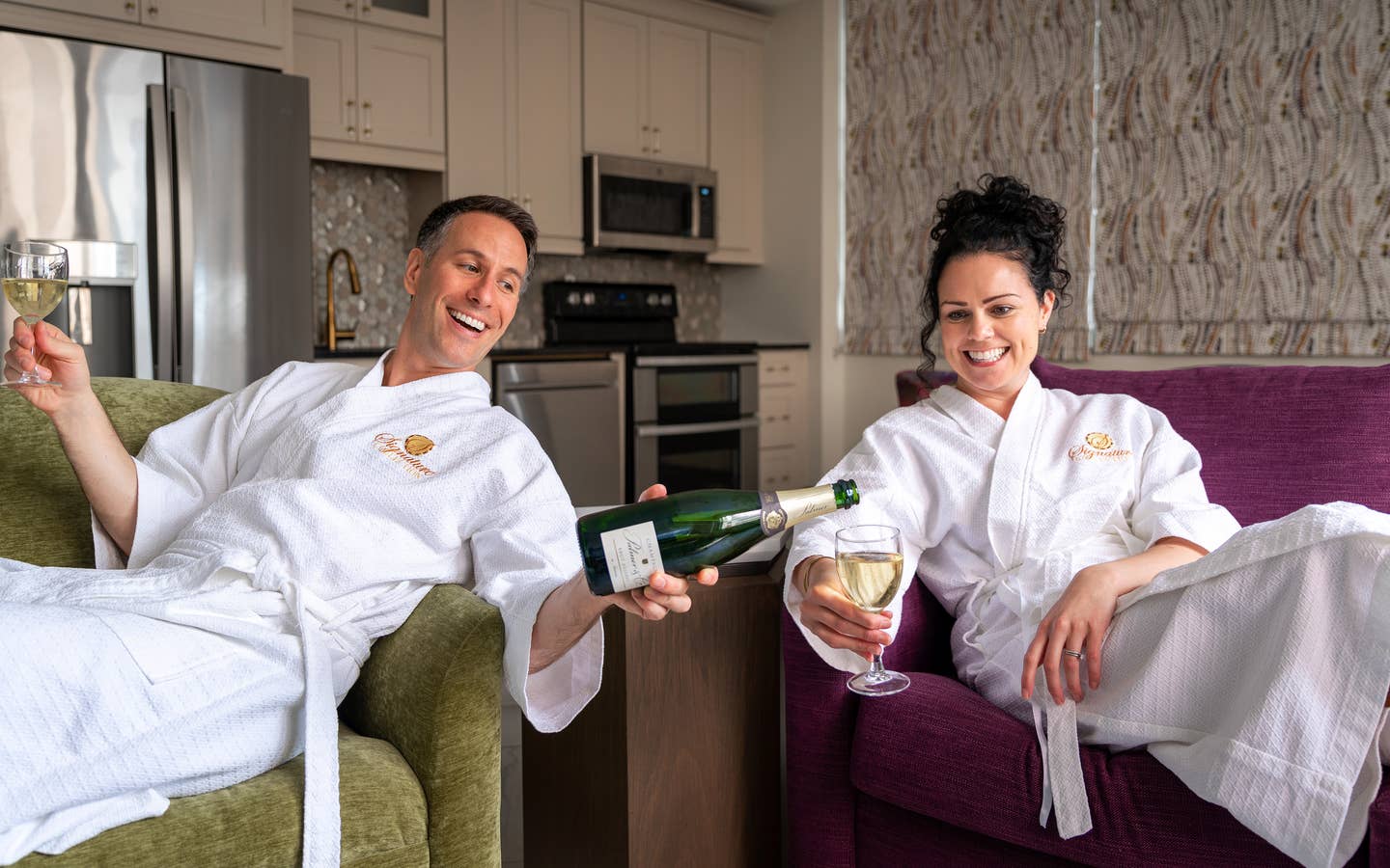 A man wearing a white robe on a green couch (left) holds a glass of champagne in his right hand while pouring the green bottle in this left hand for a woman (right) also wearing a white robe on a purple couch.
