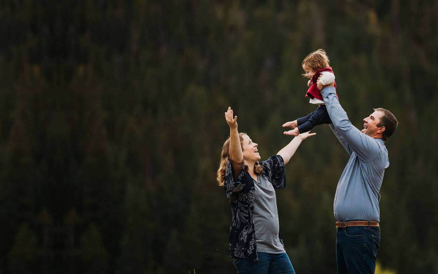 A mother (left) lifts her expressive arms as a father (right) holds up their child in front of a wooded area.