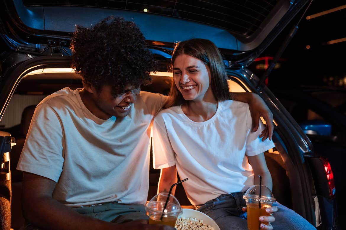 Couple at drive-in sitting in a hatchback with food and beverages.