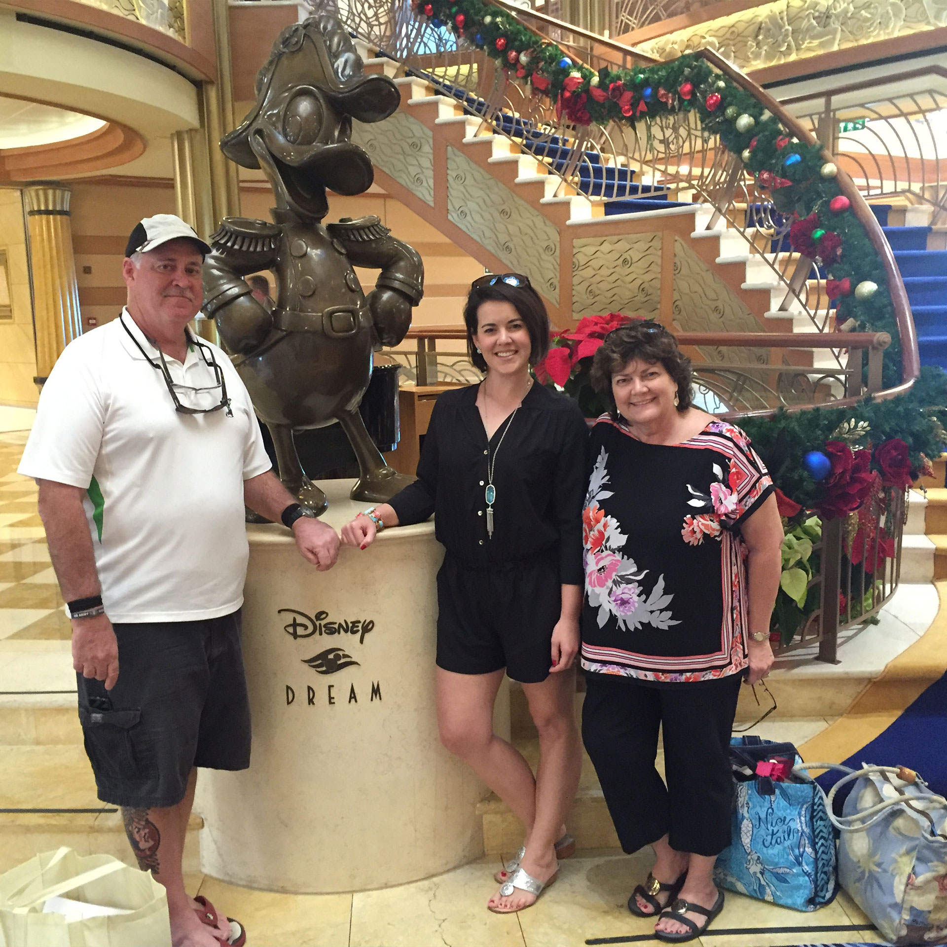 A man wearing a white and black cap, white polo and black shorts stands to the left of two women - both wearing black blouses and pants - and a bronze statue of Donald Duck on a pedestal that reads, 'Disney Dream.'