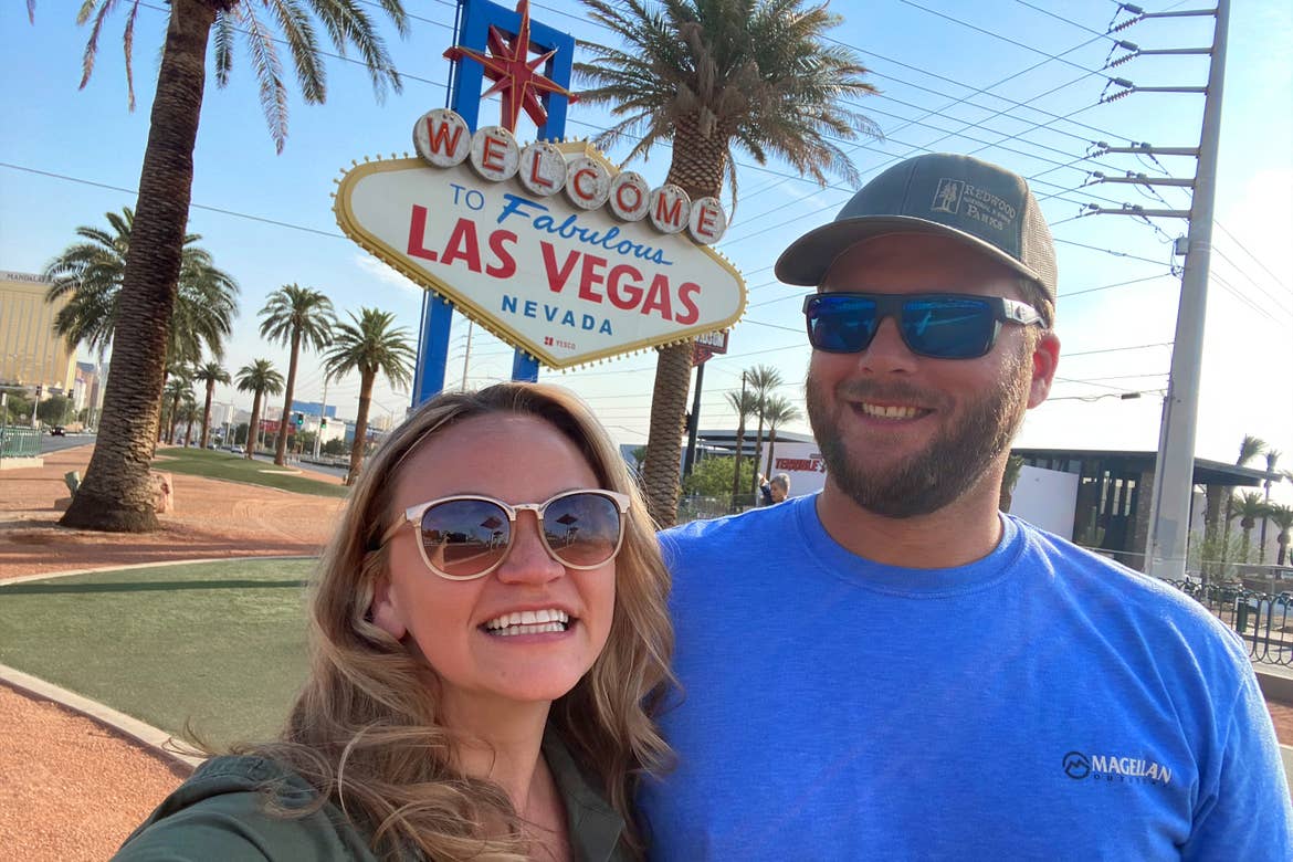 A woman wearing sunglasses (left) poses near a man wearing sunglasses and a hat (right) in front of the 'Welcome to Las Vegas Nevada' marquee sign.