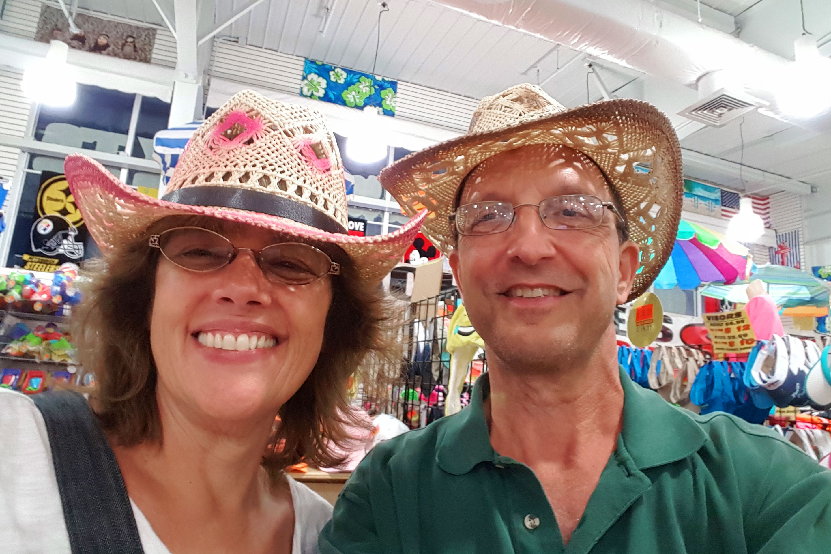A woman and man stand in a thrift store wearing straw, colored cowboy hats.