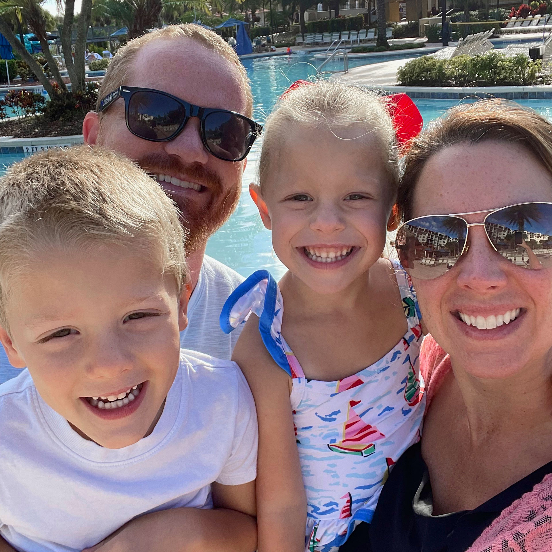 A man, woman, boy and girl wear swimsuits and trunks near an outdoor poolside.