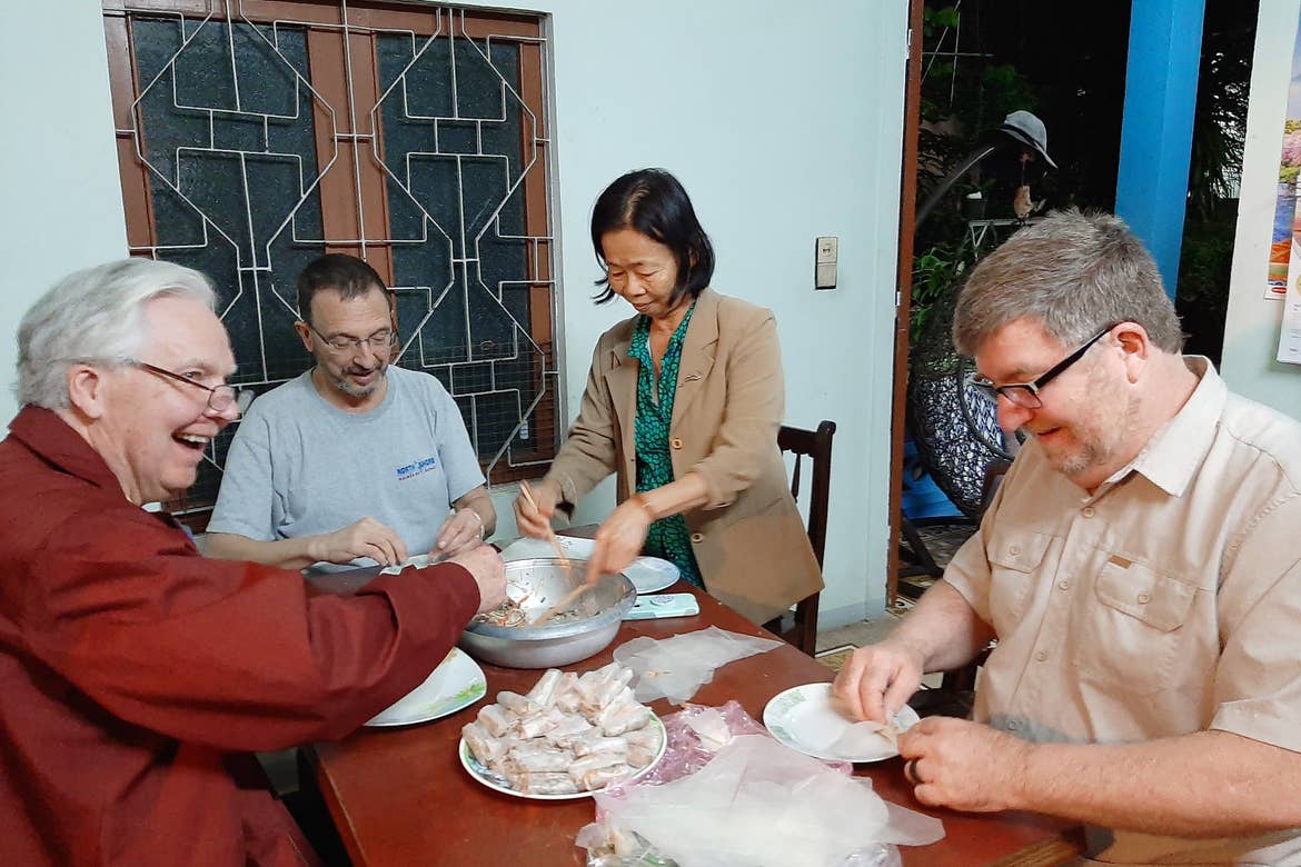 Three caucasian men (front-left, front-right, and back-left) sit at a table as a Vietnamese woman serves a traditional dish.