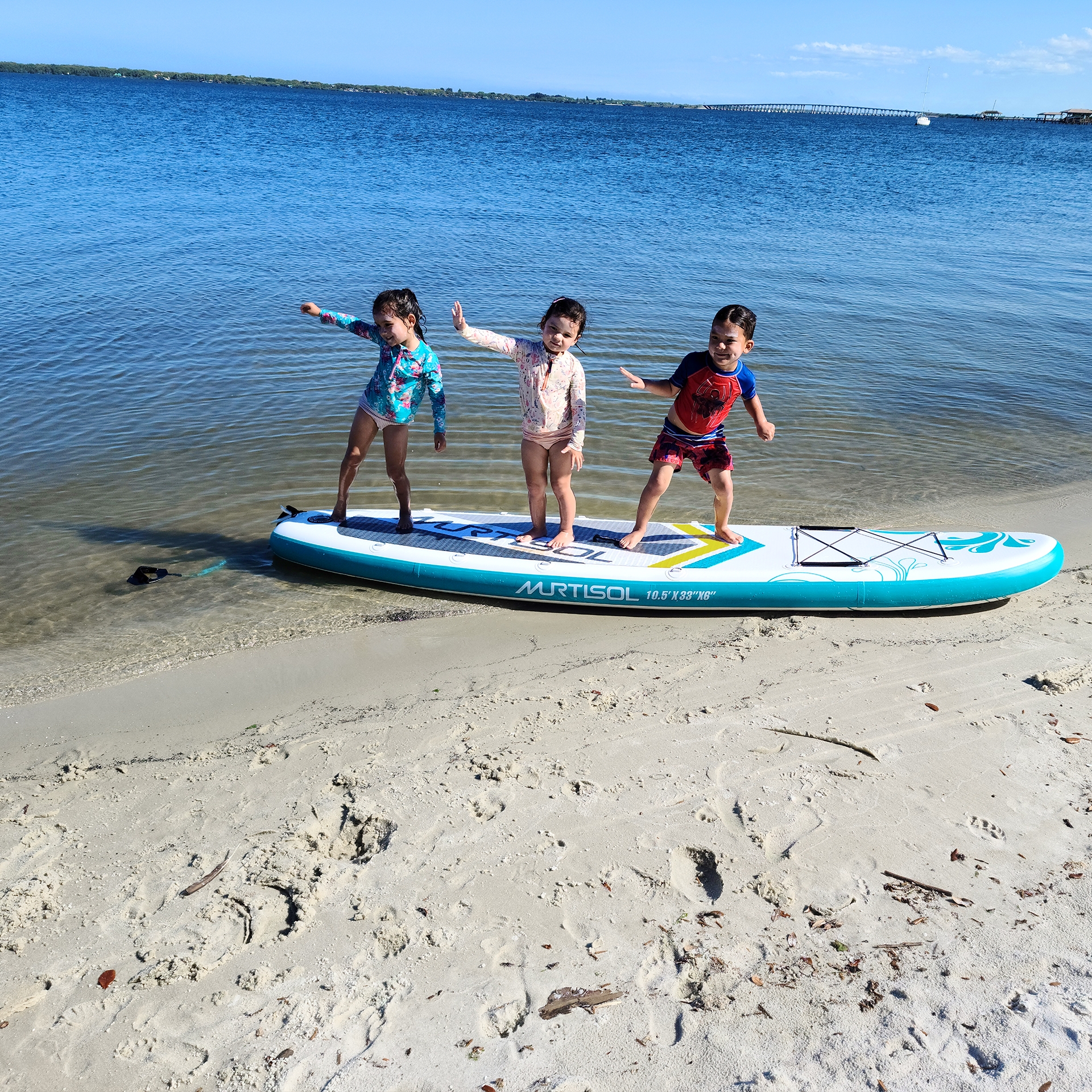 Three Asian Pacific Islander toddlers (left to right: Two girls and a boy) wear pink and blue swimsuits while standing on a paddle board on the sands and water of a beach.