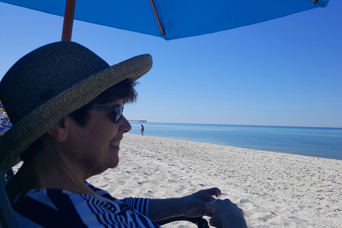 A woman in a sunhat sits under a blue umbrella on the sands of a beach under a blue sky.