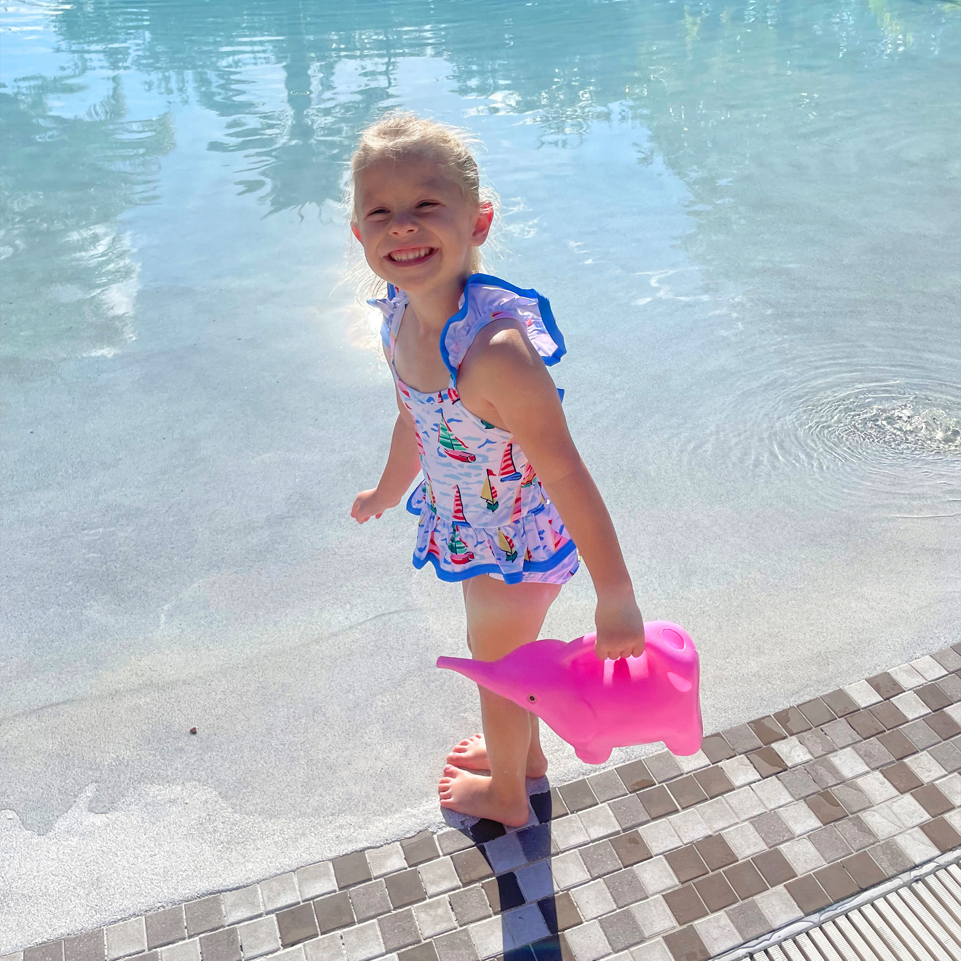A young girl wears a swimsuit and holds a pink watering can with her toes in a zero-entry pool.