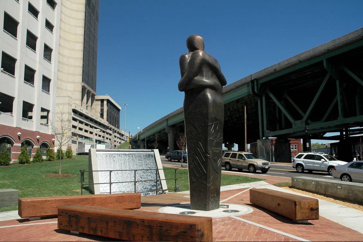 A statue made of dark, black material portrays two bodies entwined and embracing near a cityscape and bridge as cars pass on the right side.