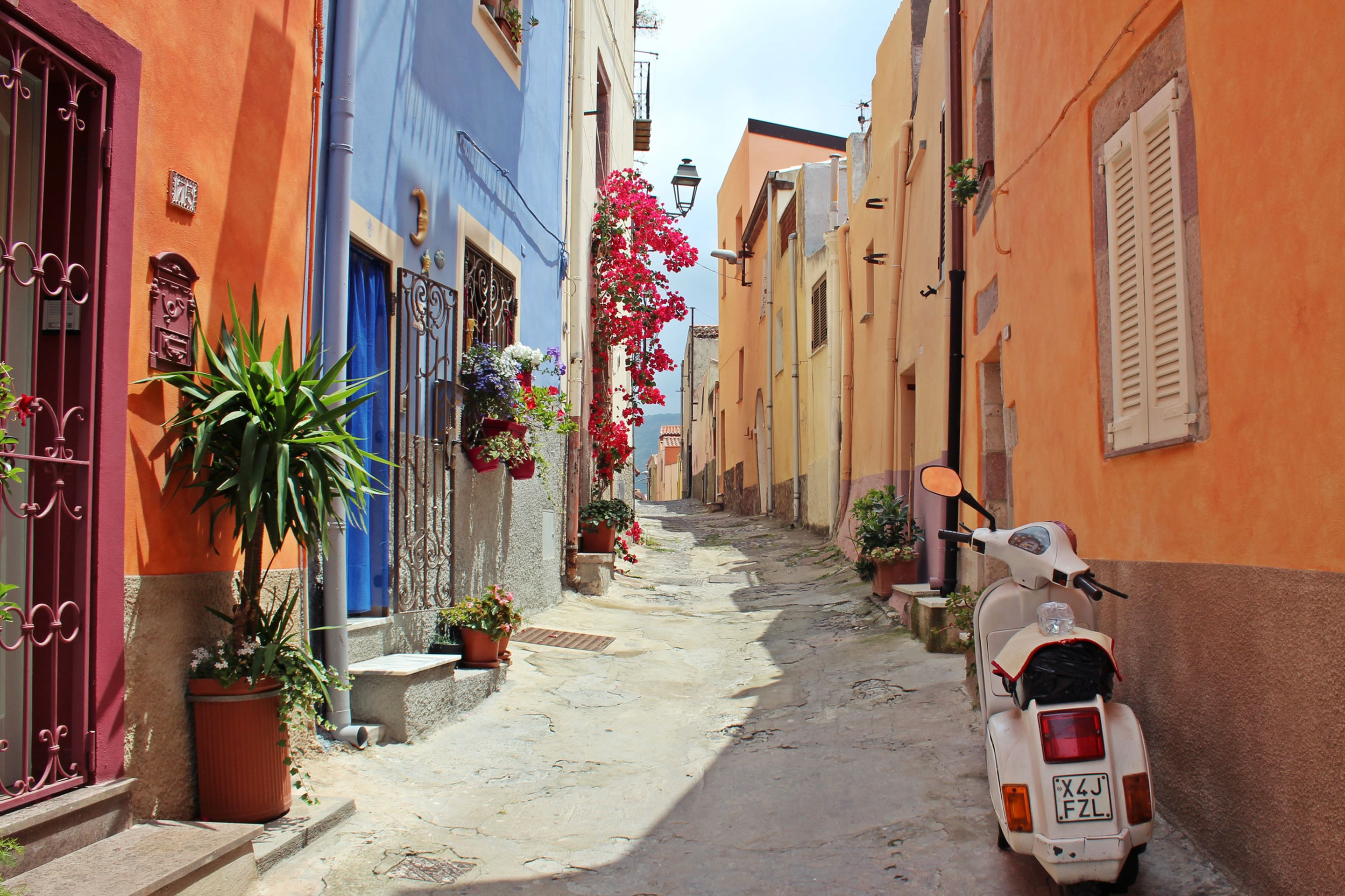 A row of colorful houses lines an alley with various flora and a white moped along each facade in Verona, Itay.